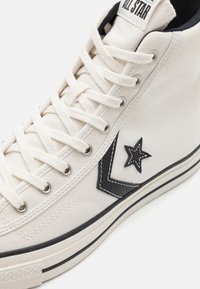 Białe sneakersy typu high-top z czarnym logo gwiazdy, białymi sznurówkami i czarną paskowaną podeszwą, pokazujące detale z góry i z boku na białym tle.