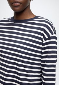 Hanorac cu dungi în bleumarin și alb. Prezintă un decolteu rotund, mâneci lungi și o croială lejeră. Material moale cu detalii vizibile la cusături.