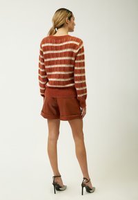 Übergroßer Pullover mit orange- und cremefarbenen Streifen und einer flauschigen Textur, kombiniert mit rostfarbenen Leder-Shorts und schwarzen High-Heels.