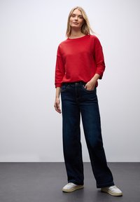 Maglietta rossa a maniche lunghe con scollatura a girocollo, abbinata a jeans a gamba larga blu scuro. Modello che indossa sneakers bianche, in piedi su uno sfondo neutro.