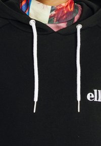 Svart hoodie med dragsnöre och vita ändar samt en logotyp. Materialet är mjukt, har en slät yta och en avslappnad passform.