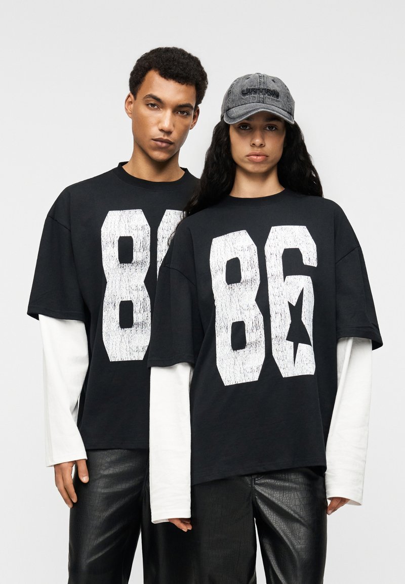 Μαύρα υπερμεγέθη t-shirts με λευκό γραφικό εκτύπωση "86", φορεμένα πάνω από λευκά μακρυμάνικα. Το μοντέλο φοράει γκρι καπέλο και γυαλιστερό μαύρο παντελόνι.