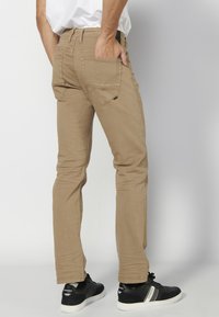 Pantalones ajustados color beige con bolsillos traseros, presillas para cinturón y costuras en contraste, combinados con zapatillas negras de suela blanca.