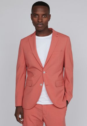 Korallenfarbener Blazer aus glattem Stoff, mit einem Ein-Knopf-Verschluss, Reverskragen, zwei vorderen Taschen und passender Hose. Darunter ein weißes T-Shirt.