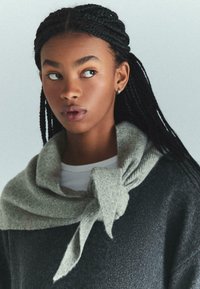 Pulover gri închis, fabricat dintr-un material moale, av�ând o eșarfă tricotată de culoare gri deschis legată la gât. Modelul are părul împletit și poartă un top alb.