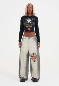 Frau mit blonden Haaren trägt ein schwarzes langärmeliges Crop-Top mit floralem Herzdesign und weite graue Hosen mit einem Kreuz- und Rosen-Patch.