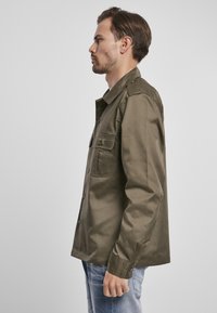Camisa verde oliva de botones fabricada en tejido suave, con dos bolsillos en el pecho y un corte relajado, combinada con unos jeans de mezclilla azul claro.