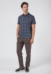Camisa de manga corta abotonada en azul oscuro con estampado floral; combinada con pantalones en marrón claro y zapatillas negras. Tela suave, corte relajado.