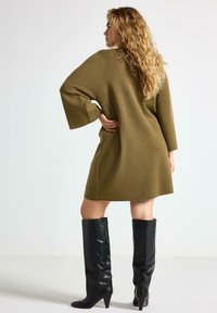 Olivenfarvet oversized sweaterkjole med lange ærmer, struktureret strikkemateriale og lige kant, parret med knæhøje sorte læderstøvler.