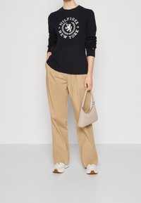 Pull côtelé bleu marine avec un logo blanc, pantalon plissé beige, petit sac à main beige assorti et baskets blanches à semelles marron clair.