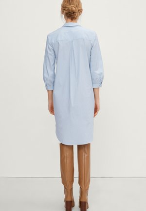 Femme aux cheveux blonds attachés en chignon, portant une chemise longue rayée bleu clair et de hautes bottes en cuir marron, debout de dos sur un fond uni.