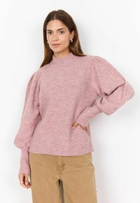 Hellrosa geripptes Sweatshirt mit hohem Kragen und puffigen Ärmeln. Der Stoff zeigt eine weiche Textur; kombiniert mit beigen Hosen und Händen in den Taschen.