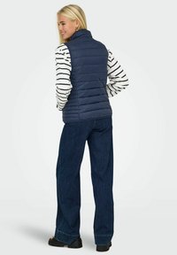 Marineblauwe puffer gilet met verticale gestikte ontwerpen, hoge kraag, gedragen over een zwart-wit gestreept shirt, gecombineerd met wijde denim jeans.
