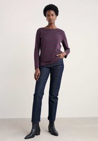 Frau steht da und trägt ein lila und schwarz gestreiftes Langarmshirt, dunkelblaue Jeans, schwarze Stiefeletten und goldene Creolen.