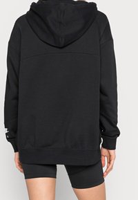 Person som bär en svart överdimensionerad huvtröja med huvan nedfälld och svarta cykelshorts, visad bakifrån mot en enfärgad bakgrund.