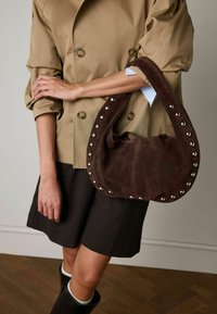 Borsa in camoscio marrone con manico curvo, arricchita da borchie argentate lungo la cucitura. Indossata con una giacca beige e pantaloncini a righe sottili.