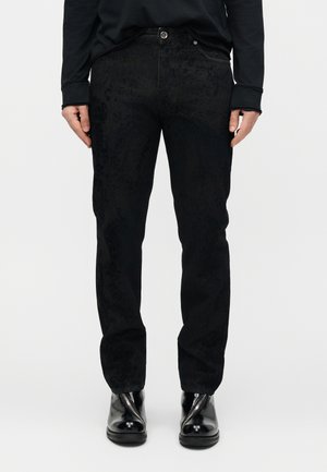 Persona che indossa pantaloni neri dritti con trama, top nero a maniche lunghe e stivaletti neri lucidi, in piedi su uno sfondo bianco semplice.