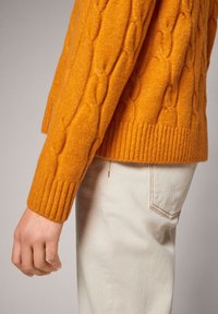 Maglione arancione a maglia rasata con orlo a coste, dotato di maniche lunghe e un motivo testurizzato, abbinato a pantaloni chiari.