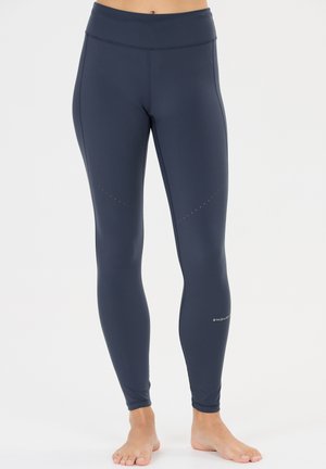 Nærbillede af en person iført mørkeblå tætsiddende atletikleggings med høj talje og lille reflekterende logo nær anklen.