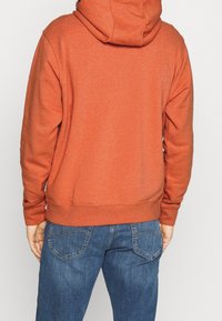 Rostorange hoodie med dragsko i huvan, ribbade ärmslut och nederkant. Bärs med blå denimjeans som har synliga bakfickor.