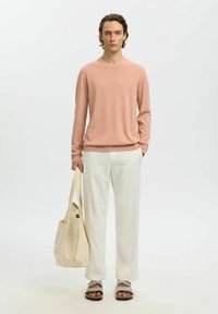Jeune homme debout, portant un pull rose clair, un pantalon blanc, des sandales beiges, et tenant un sac fourre-tout beige dans un décor blanc minimaliste.