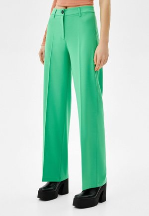 Femme portant un pantalon large taille haute vert vif avec pli frontal, des chaussures à plateforme noires à talons, et un haut court pêche à manches longues.