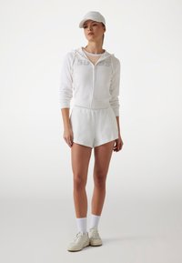 Weißer Zip-Hoodie mit geprägtem Logo, kombiniert mit weißen Shorts. Accessoires umfassen eine weiße Mütze und weiße Sneaker. Strukturierter Stoff, lässiges Design.