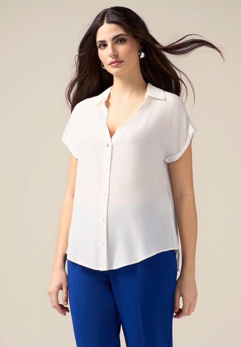 Witte blouse met korte mouwen en knoopsluiting, gemaakt van een zachte stof en met een licht gebogen onderrand, gecombineerd met blauwe broek.