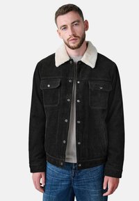 Chaqueta de pana negra con un collar de sherpa blanco; presenta bolsillos en el pecho y botones de metal. Combinada con una camisa gris claro y jeans azules.