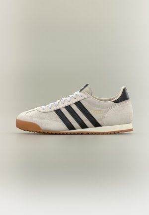 Hvid Adidas sneaker med sorte striber, gummisål og gulddetalje med logo, vist i sideprofil på en neutral baggrund.