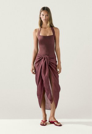 Woman wearing sleeveless maroon dress with a knotted front slit, standing barefoot against a plain light background.