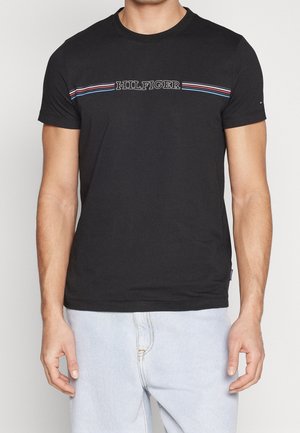 Mężczyzna w czarnym T-shirtcie z napisem "HILFIGER" i czerwono-biało-niebieskimi poziomymi paskami na wysokości klatki piersiowej, w połączeniu z jasnoniebieskimi dżinsami.
