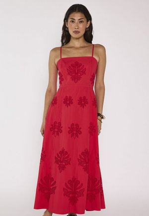 Femme portant une robe rouge sans manches, longueur cheville, à fines bretelles et motifs floraux brodés, debout sur un fond blanc uni.
