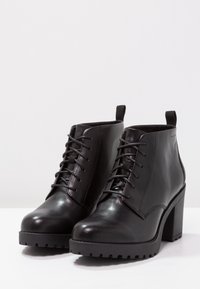 Botas de cuero negro hasta el tobillo con un alto tacón cuadrado, punta redondeada, cordones en la parte delantera y suela de goma texturizada. Presentan detalles de costura y una lengüeta para facilitar su uso.