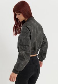 Črna skrajšana bomber jakna iz gladkega, teksturiranega blaga z zadrgo spredaj, rebrastimi manšetami in stranskim žepom. Model nosi temne kavbojke.