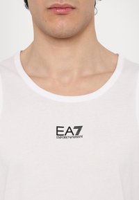 Balta medvilninė marškinėlė su apykakle ir juodu EA7 Emporio Armani logotipu spausdintu ant krūtinės. Lygus paviršius, aptemptas dizainas.