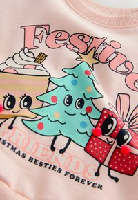 Helles rosa Sweatshirt mit Cartoon-Grafiken: Kuchen, Weihnachtsbaum und Geschenk, mit kräftigen Farben und Pom-Pom-Akzenten. Der Text lautet "Festliche Freunde."