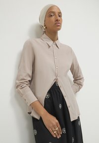 Camicia button-up beige chiaro in tessuto texturizzato, con maniche lunghe e orlo curvo, abbinata a una gonna nera con motivo floreale.