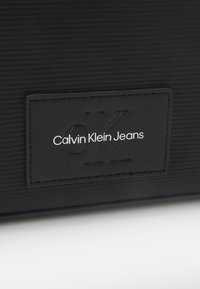 Sac en similicuir noir avec une surface texturée, présentant une étiquette embossée portant l'inscription "Calvin Klein Jeans" en lettres blanches et noires.