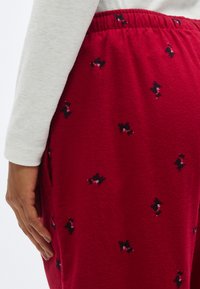 Pantalon de pyjama en polaire rouge avec un motif de chiens en noir et blanc. La ceinture est élastique et froncée pour le confort. La texture est douce.