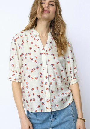 Femme portant un chemisier blanc à boutons avec de petits motifs floraux rouges et violets, manches trois-quarts, associé à un jean bleu et un bracelet.