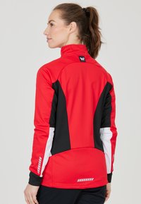 Giacca atletica rossa e nera con colletto alto, tessuto testurizzato, dettagli bianchi a contrasto, tasche con zip e riflessi.