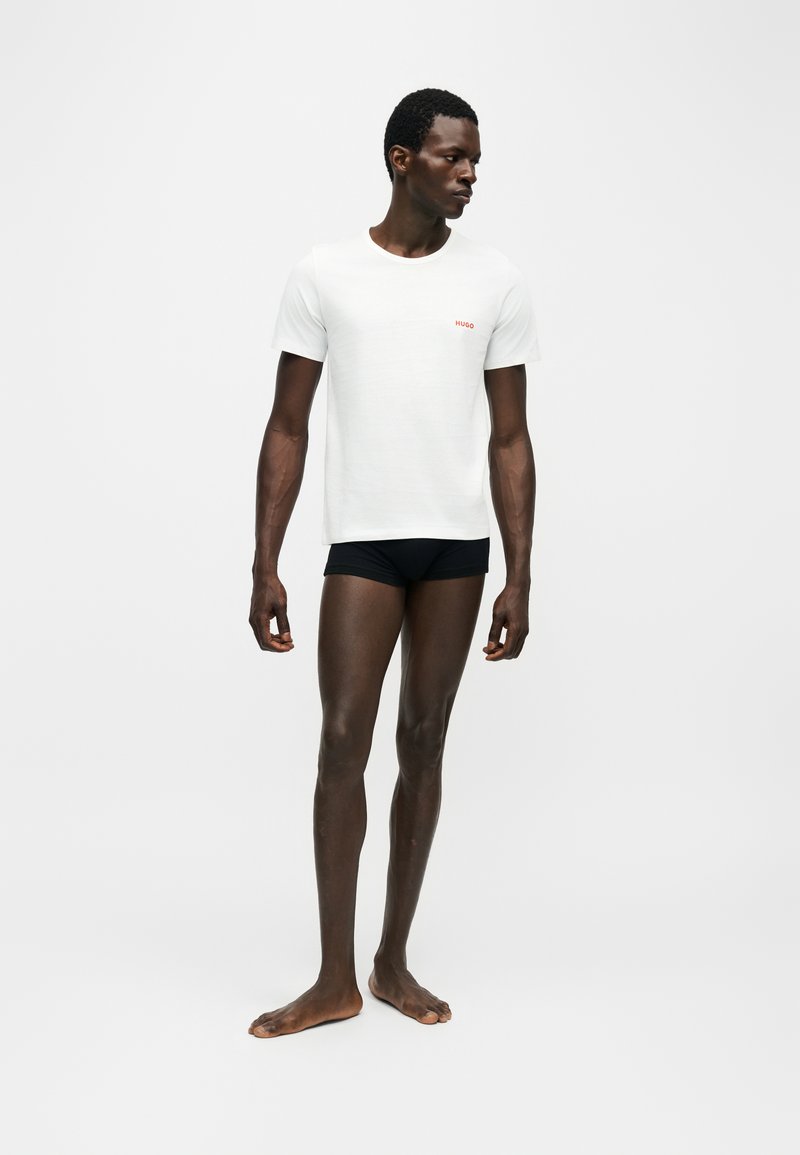Mann steht barfuß, trägt ein weißes Kurzarm-T-Shirt mit kleinem roten Text auf der Brust und schwarze enganliegende Shorts vor einfarbigem Hintergrund.