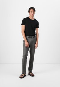 Svart t-shirt med kort ärm, grå skräddarsydda byxor och svarta sandaler med tjocka remmar. Modell står mot en enkel vit bakgrund.
