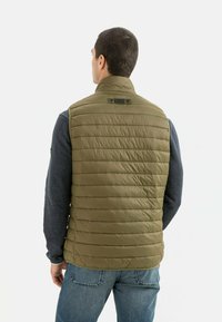 Olivgrüne, gepolsterte Weste mit vertikaler Naht, hohem Kragen und Etikett auf der Rückseite. Getragen über einem grauen Langarmshirt und blauen Jeans.