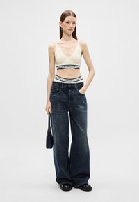 Krémový bralet s čiernym logom na pásku, skombinovaný s širokými tmavomodrými džínsami. Model drží malú čiernu tašku a má na sebe čierne topánky.