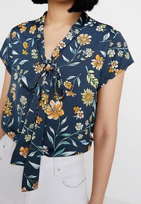 Blusa floral en azul marino con mangas cortas y un lazo en la parte delantera. Presenta vibrantes patrones florales en naranja, amarillo y verde sobre un tejido suave.