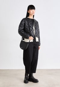 Vrouw in een zwarte gewatteerde jas, zwarte wijde broek, zwarte laarzen, met een kleine zwarte schoudertas tegen een effen achtergrond.