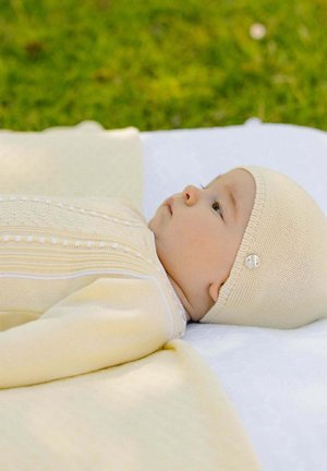 Bébé allongé sur une couverture blanche en plein air, portant une tenue en tricot jaune avec un bonnet assorti, semblant calme et regardant vers le haut.