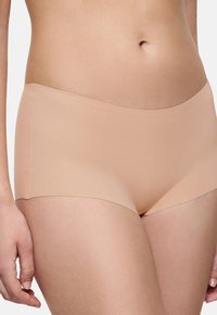 Ženska, oblečena v brezšivne spodnje hlačke nude barve, ki prikazuje srednji del telesa in zgornje stegno na enobarvni podlagi.